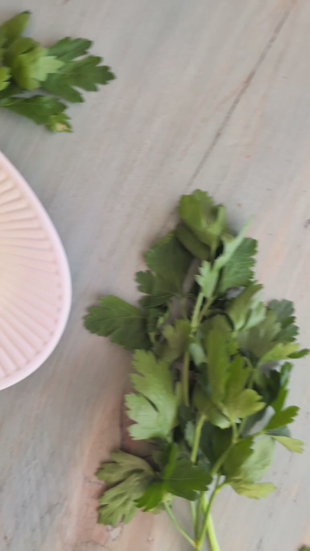 Load video: Fresh flat-leaf parsley sprigs on a pale wooden board beside a pale pink-rimmed dish
