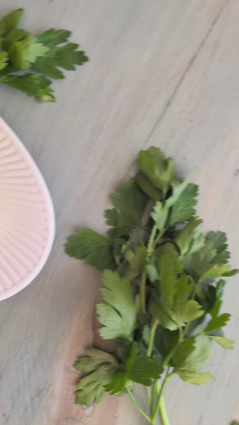 Fresh flat-leaf parsley sprigs on a pale wooden board beside a pale pink-rimmed dish