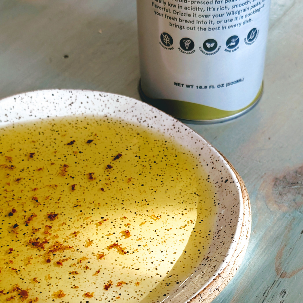 Shallow speckled bowl of olive oil with chili flakes on wood table; bottle in background labeled NET WT 16.9 FL OZ (500mL)