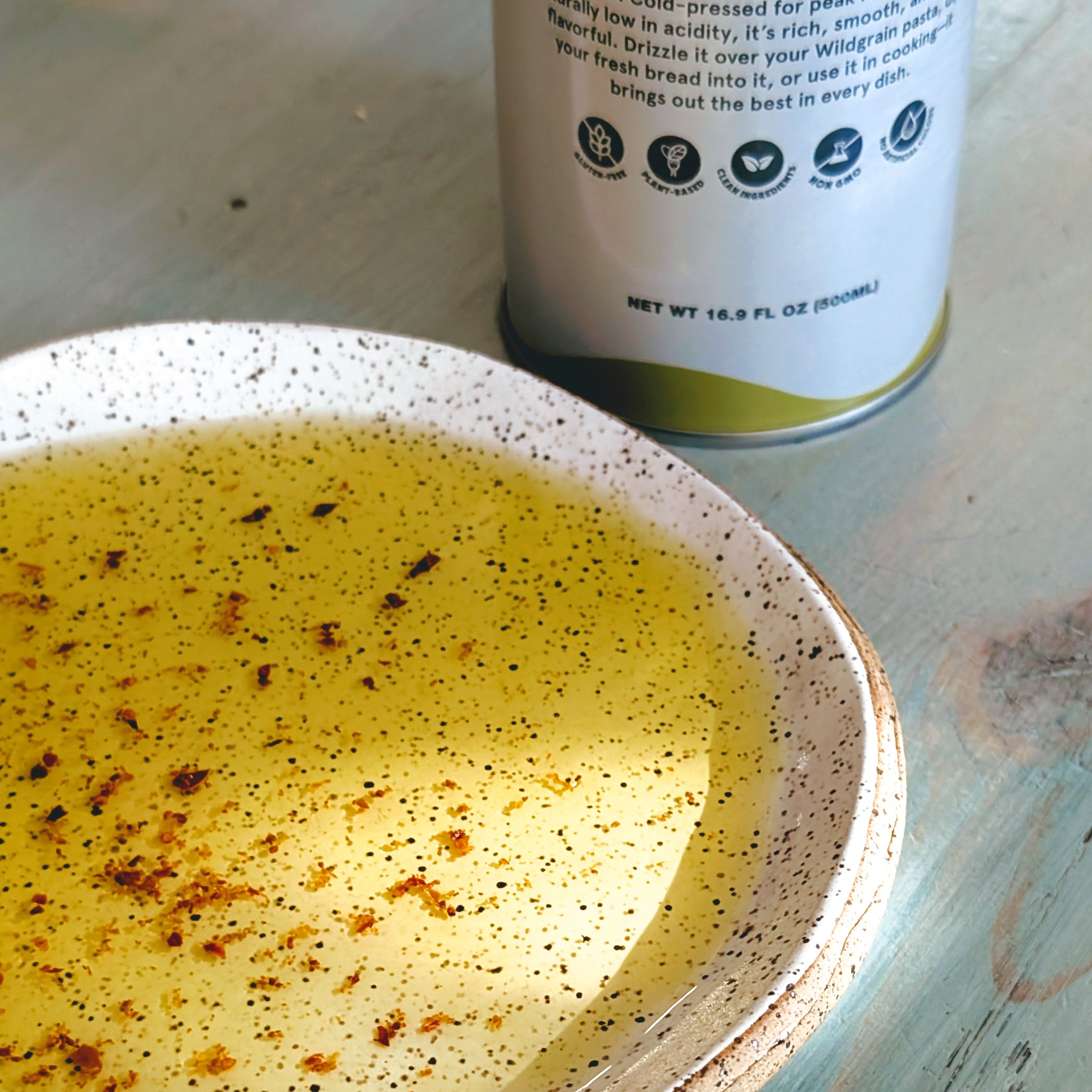 Shallow speckled bowl of olive oil with chili flakes on wood table; bottle in background labeled NET WT 16.9 FL OZ (500mL)