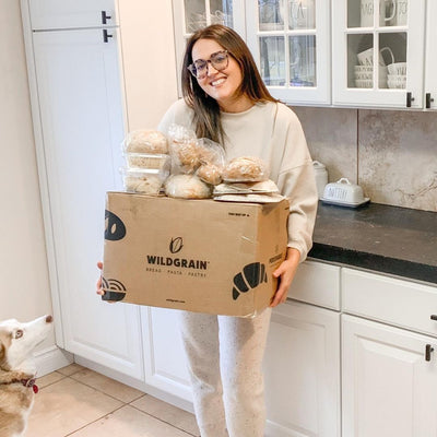 Smiling woman in a kitchen holding a Wildgrain delivery box filled with bread and pastries, a dog looks up at her.