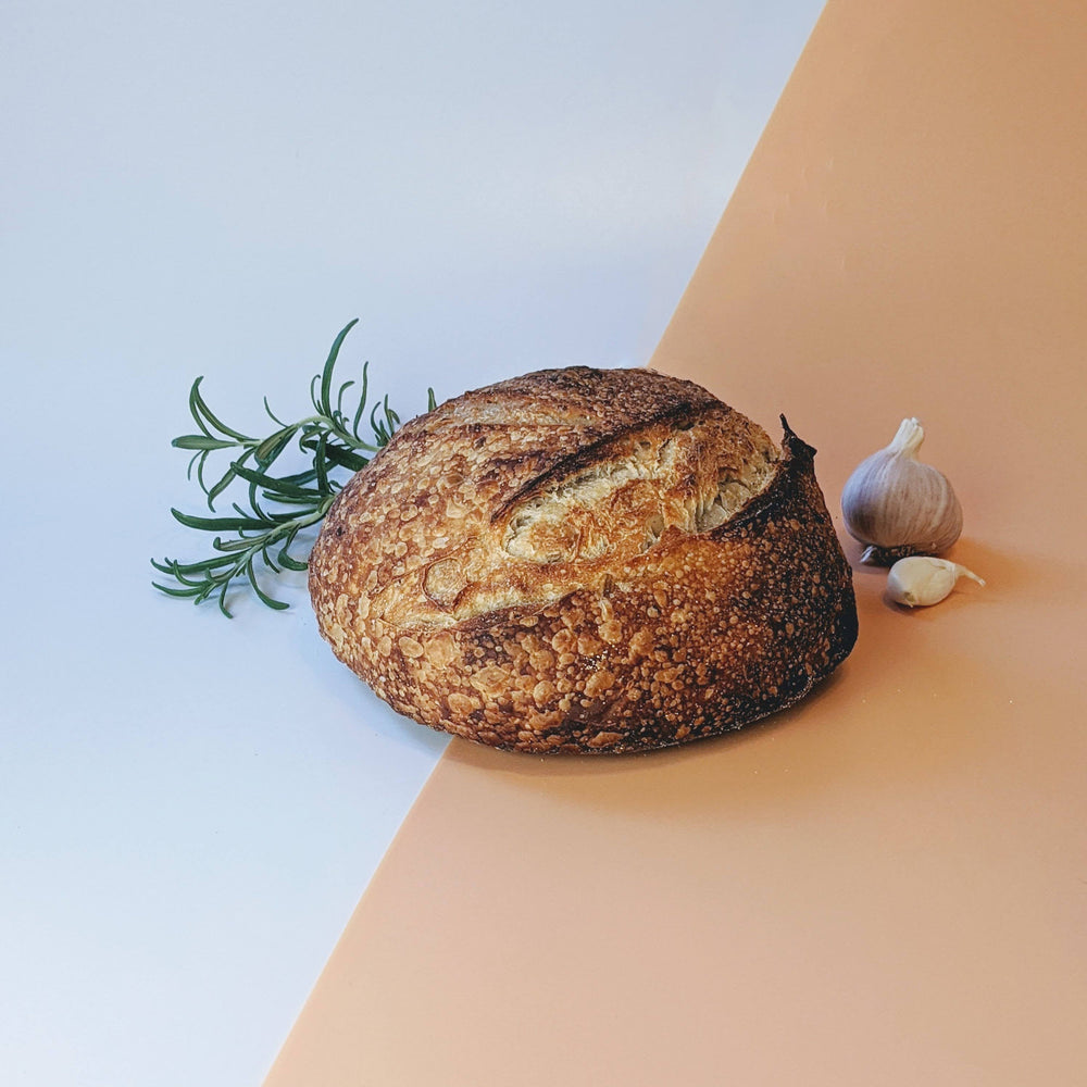 Round sourdough loaf with rosemary sprig and garlic clove on split pale blue and peach background