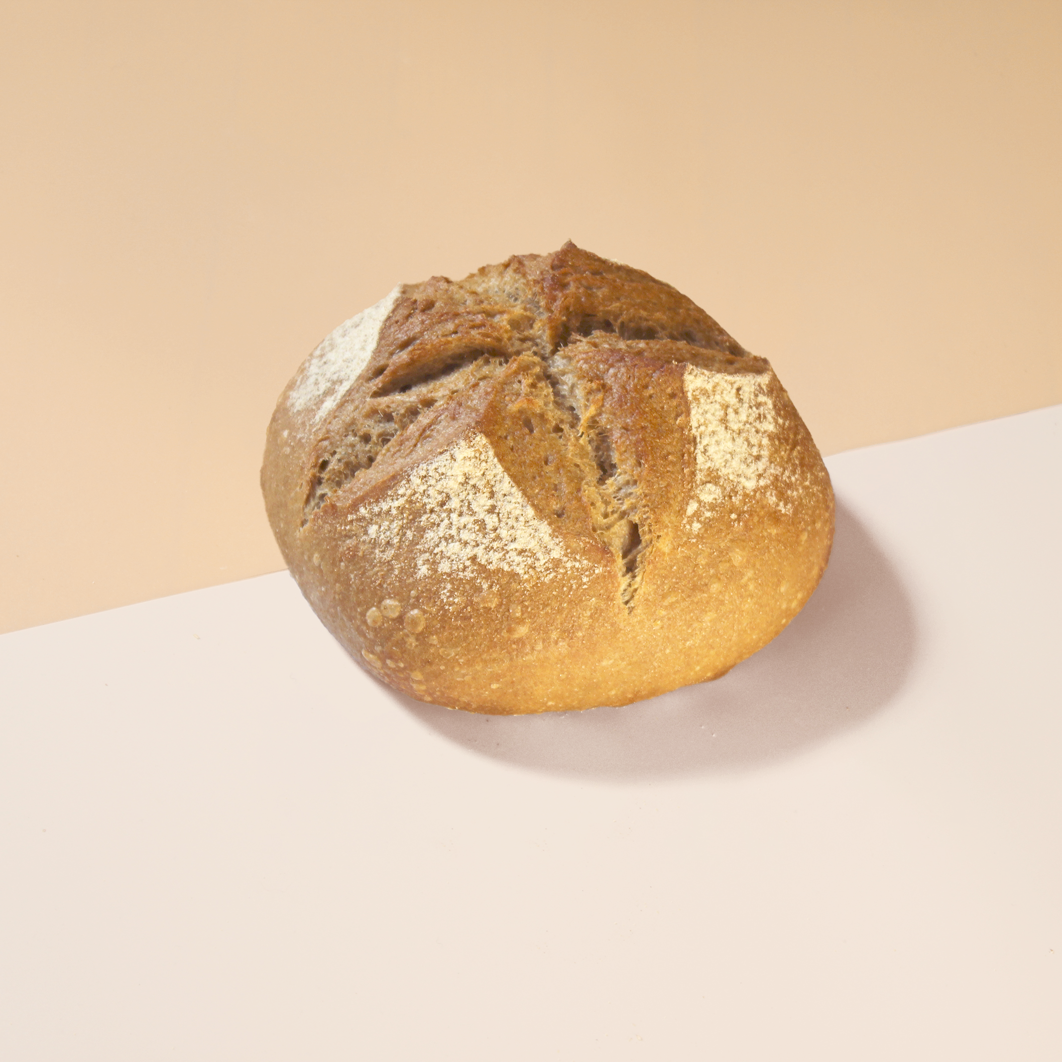 Round artisan sourdough loaf with cross scoring and flour dusting on a two-tone peach and pale-pink background.