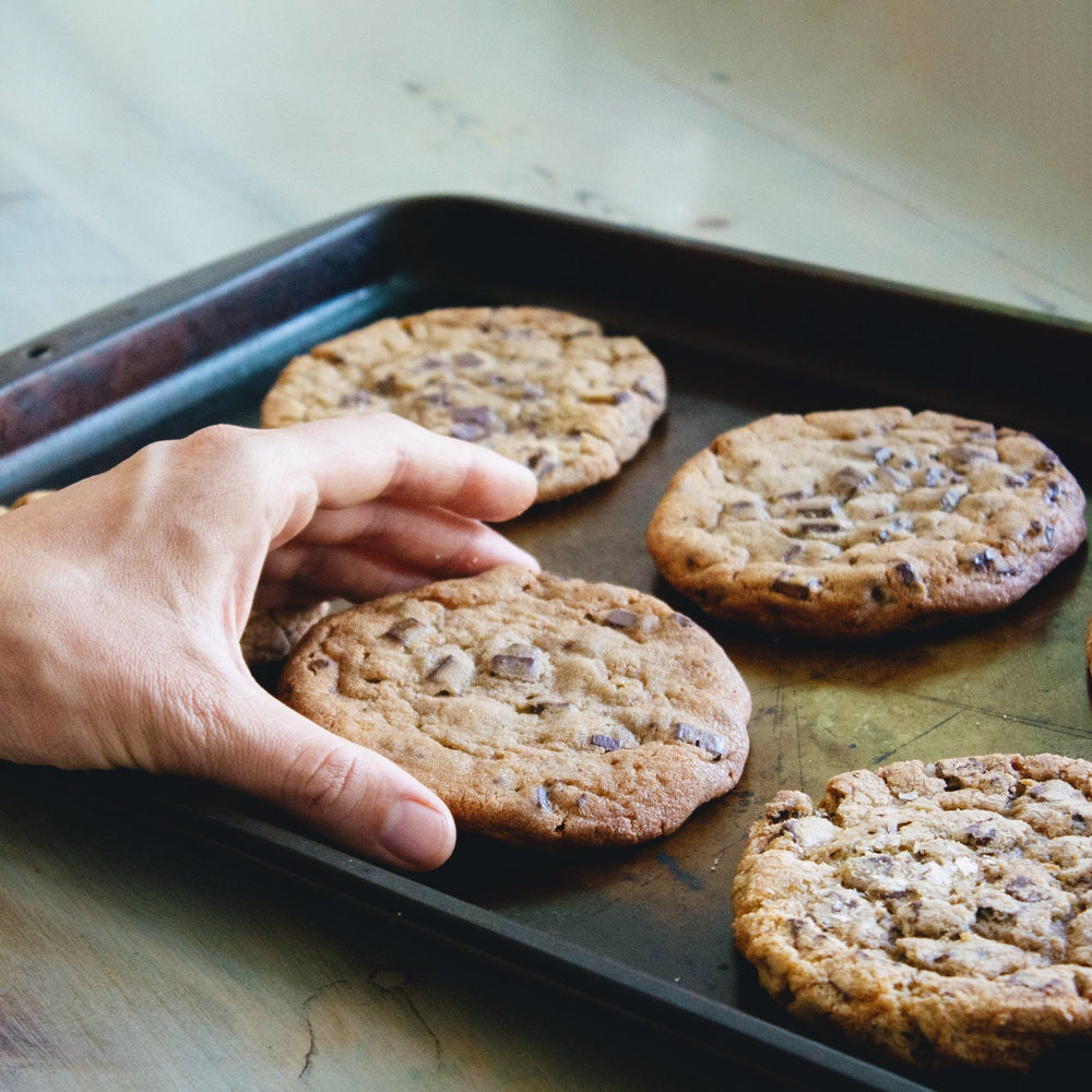 A hand reaching for a plant-based giant chocolate chip cookie on a baking tray with four cookies.