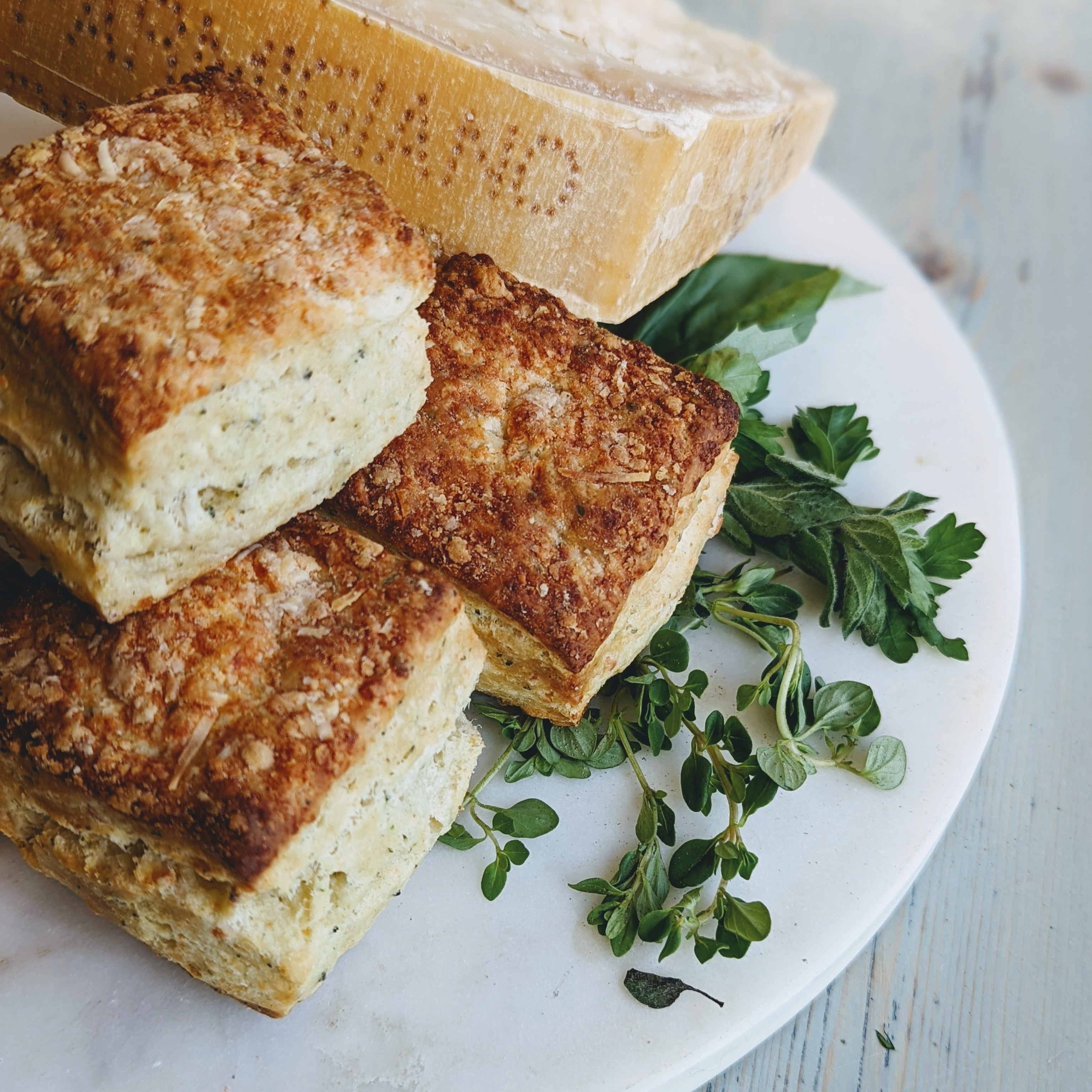 Three square Parmesan-herb biscuits on a marble plate with fresh herbs and a wedge of Parmesan cheese.