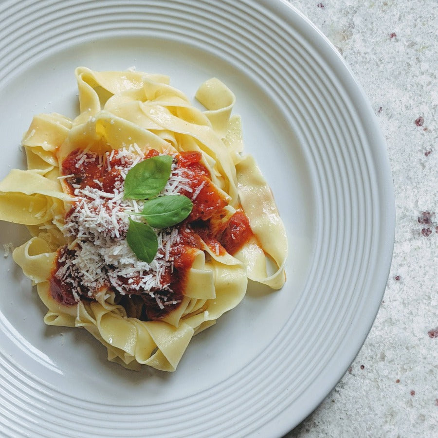 Plate of pappardelle pasta topped with tomato sauce, grated cheese and basil on a white plate
