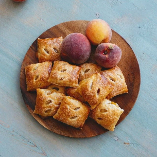 A wooden plate with peach pie bites and fresh peaches on a blue surface.