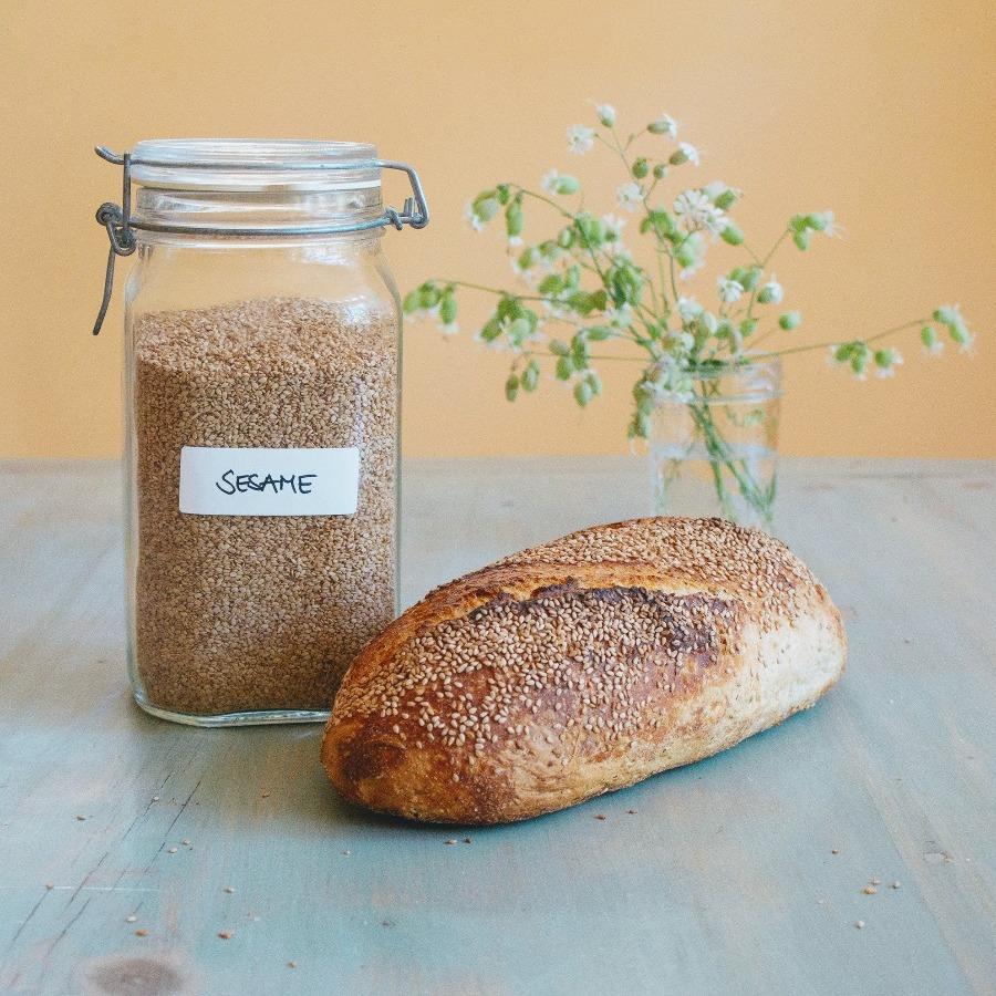 A loaf of Sourdough Sesame Seed bread next to a jar labeled 'SESAME' filled with sesame seeds, with flowers in the background.