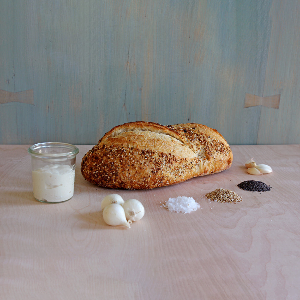 Seeded loaf on pale wood table with jar of white spread, small white bulbs, garlic cloves, and piles of salt and seeds.
