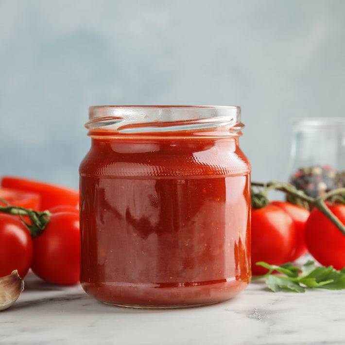 Front view of glass jar of marinara sauce showcasing smooth red sauce with tomatoes and basil in the background