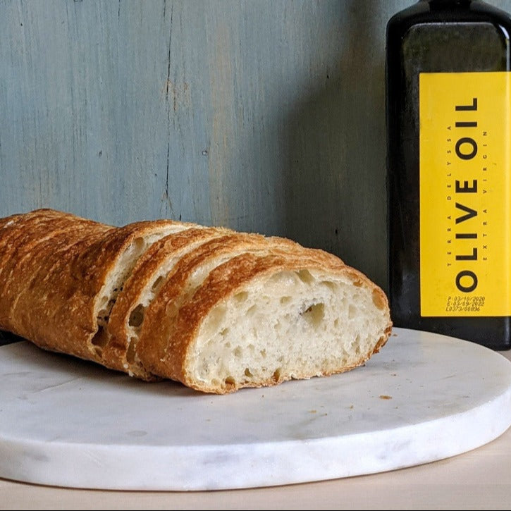 A sliced Wildgrain Olive Oil Ciabatta loaf on a marble surface next to a bottle of extra virgin olive oil.