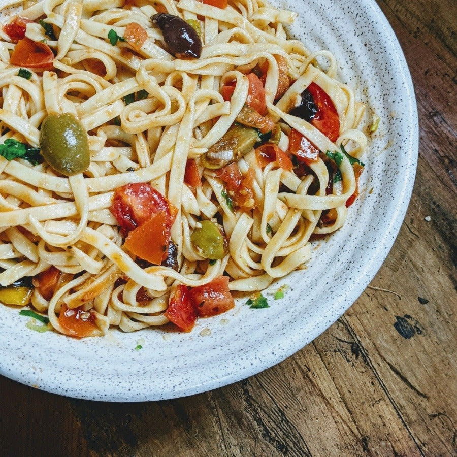 Close-up of a plate of pasta with green and black olives, diced tomatoes, and herbs on a speckled white plate on a wooden table.
