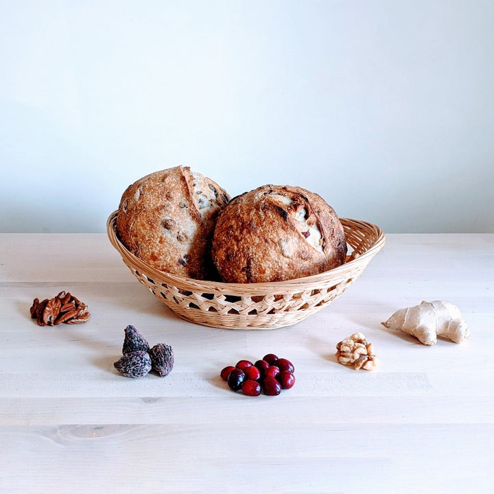 Two round rustic loaves in a woven basket on light wood table, with pecans, figs, cranberries, walnuts, and ginger.