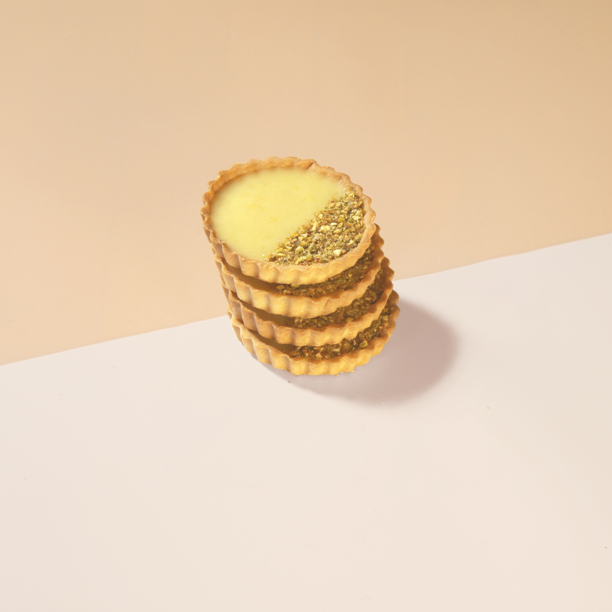 Stack of four round tarts with yellow filling and crushed nuts on one side, on a two-tone beige and white background.