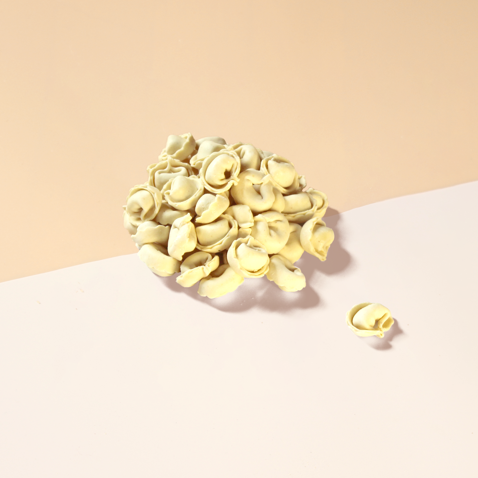 Uncooked tortellini piled on a two-tone pale peach and pink surface with one tortellino separated to the right.